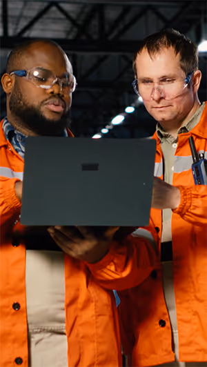 Two factory workers wearing orange safety jackets and safety glasses reviewing data on a laptop in an industrial setting.